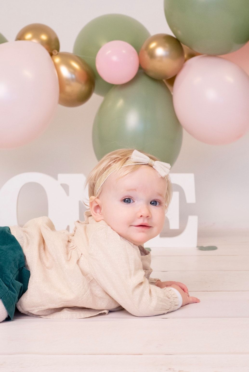 Petite fille au noeud dans les cheveux allongée sous une guirlande de ballons verts et roses, séance premier anniversaire