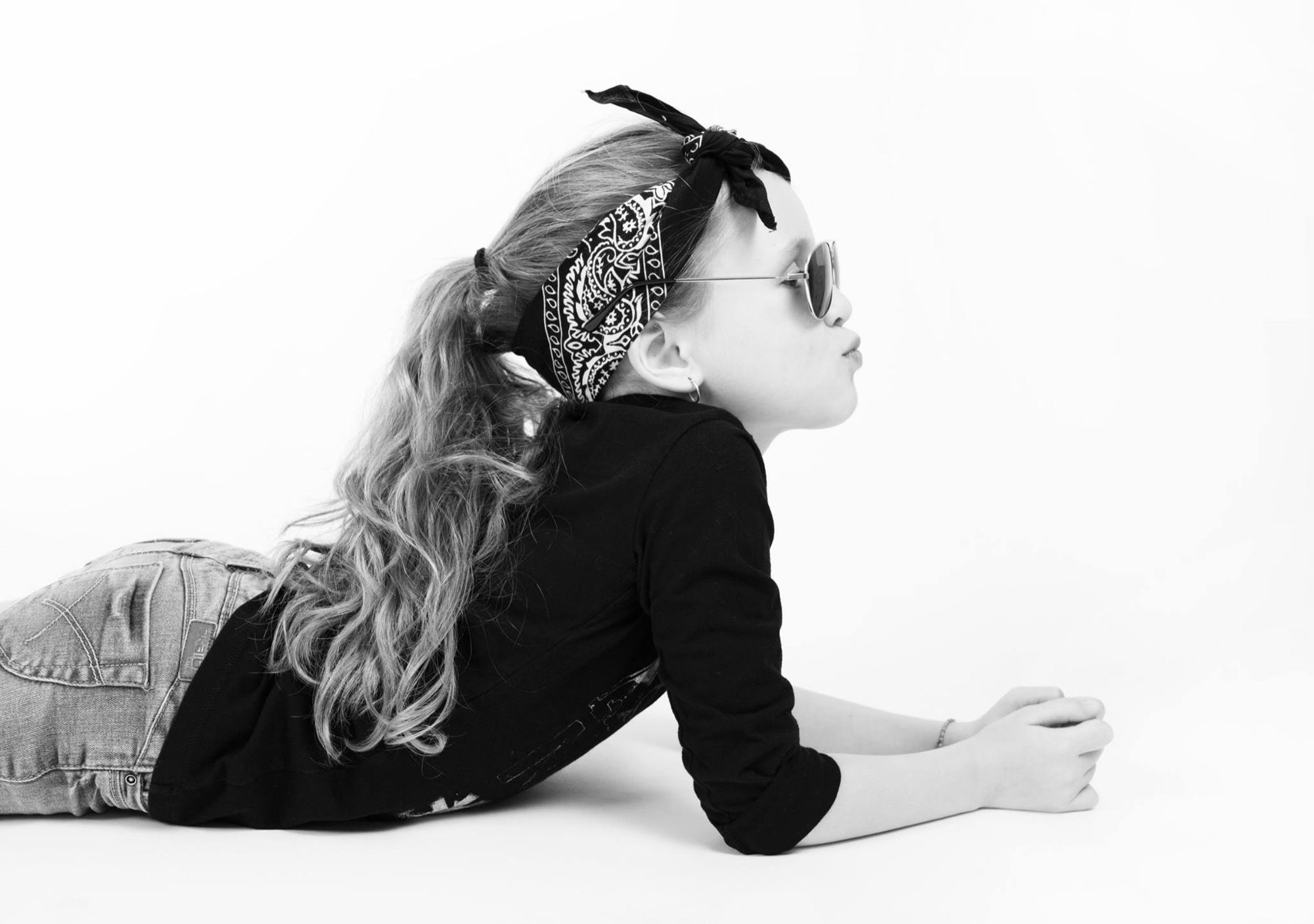 Little girl lying down with a bandana and sunglasses blowing a kiss, black and white child portrait at the studio