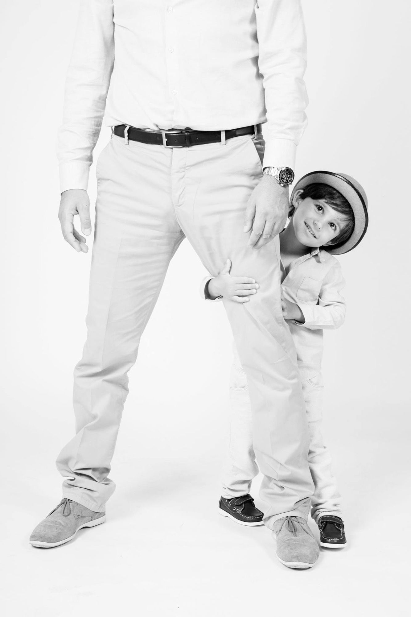 Little boy in a hat hiding behind his dad's legs, playful black and white child portrait at the studio