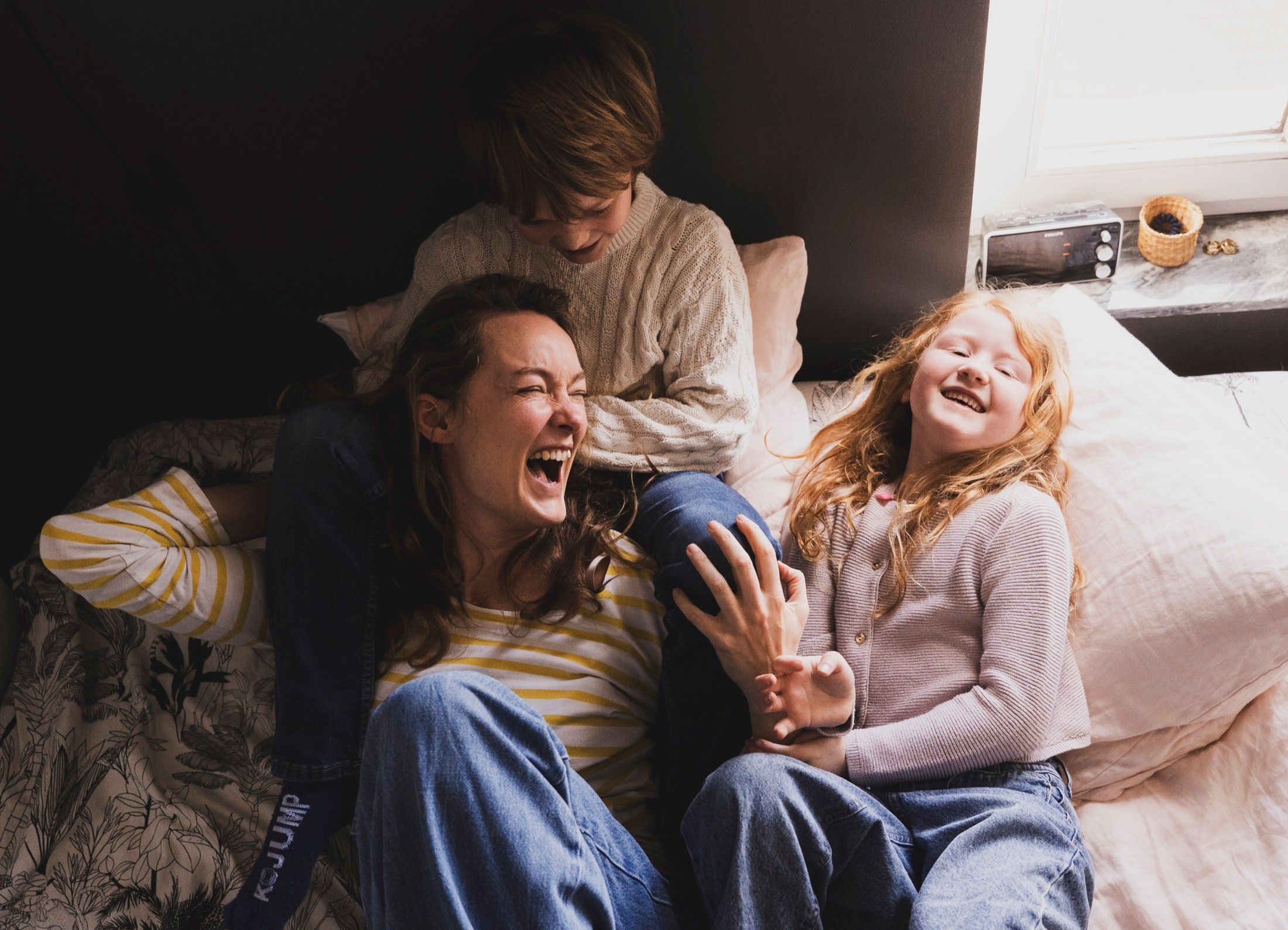 Maman riant aux éclats sur un lit avec ses deux enfants, séance famille à domicile
