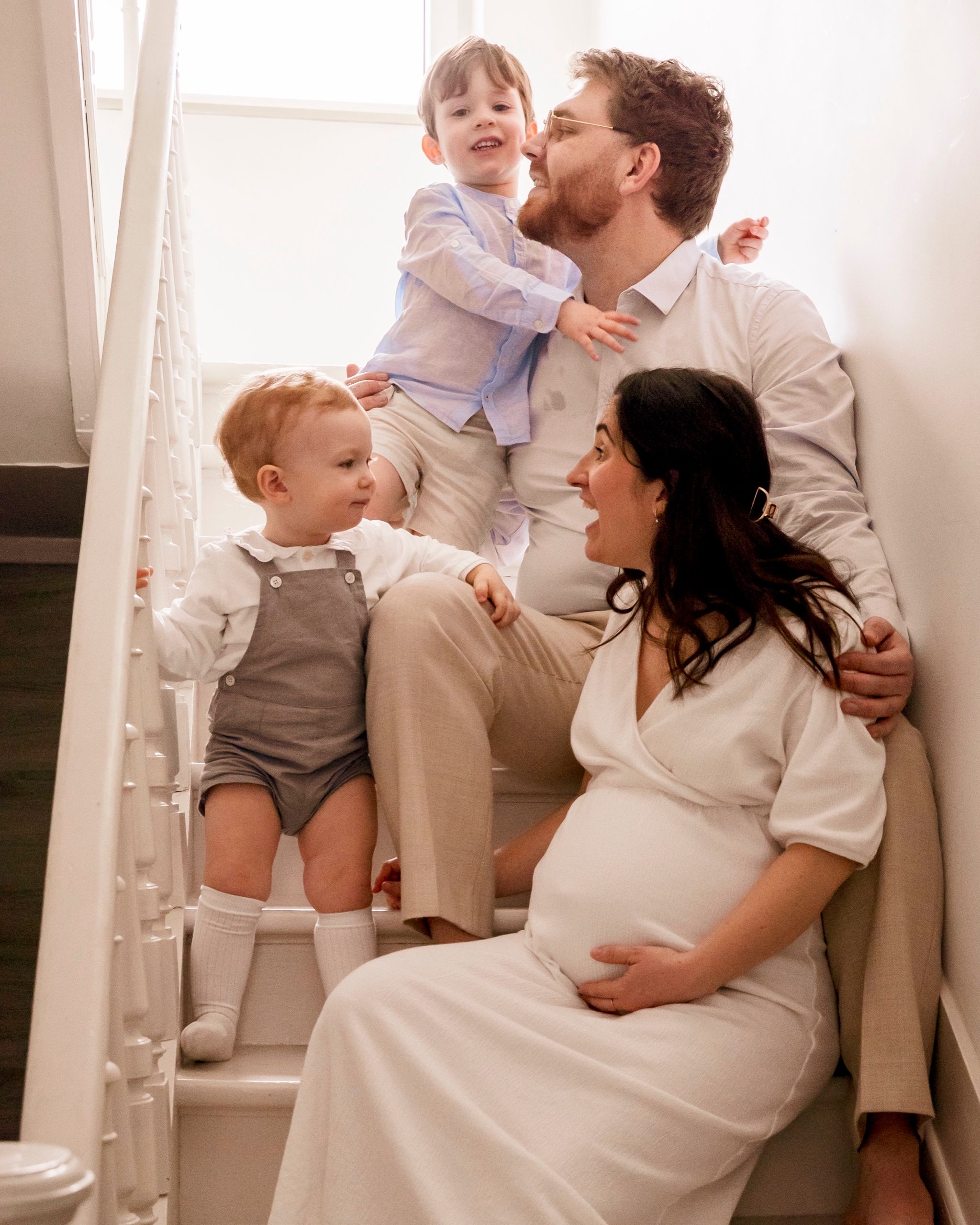 Famille dans un escalier à domicile : parents et deux jeunes enfants, mère enceinte, tenues claires et lumière douce