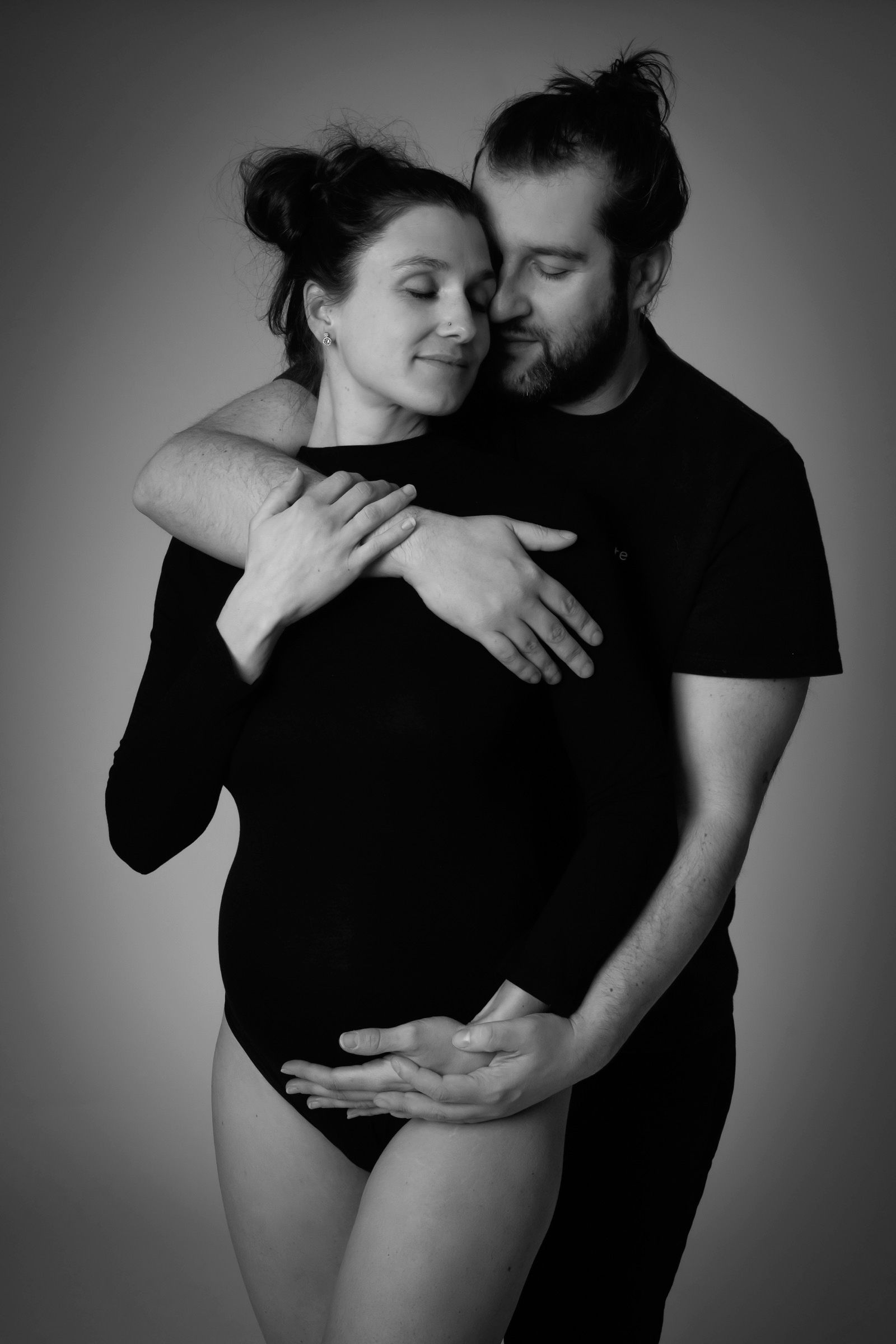 Couple enlacé en tenues noires, mains posées sur le ventre de la future maman, portrait de grossesse en noir et blanc