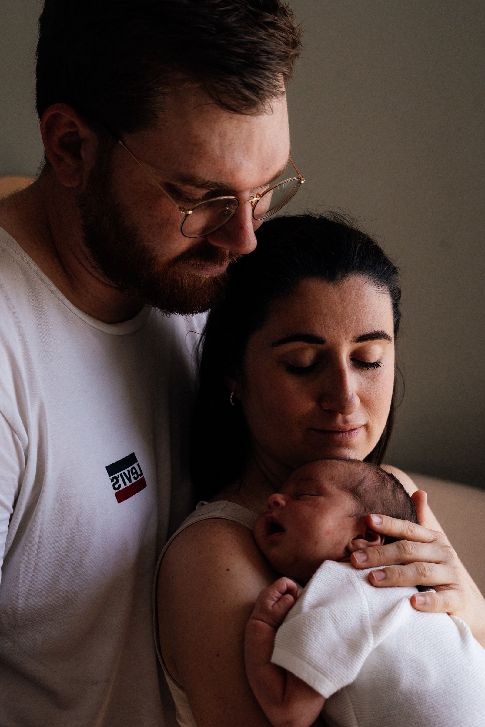 Parents tenant leur nouveau-né endormi contre eux, séance naissance à domicile, lumière douce