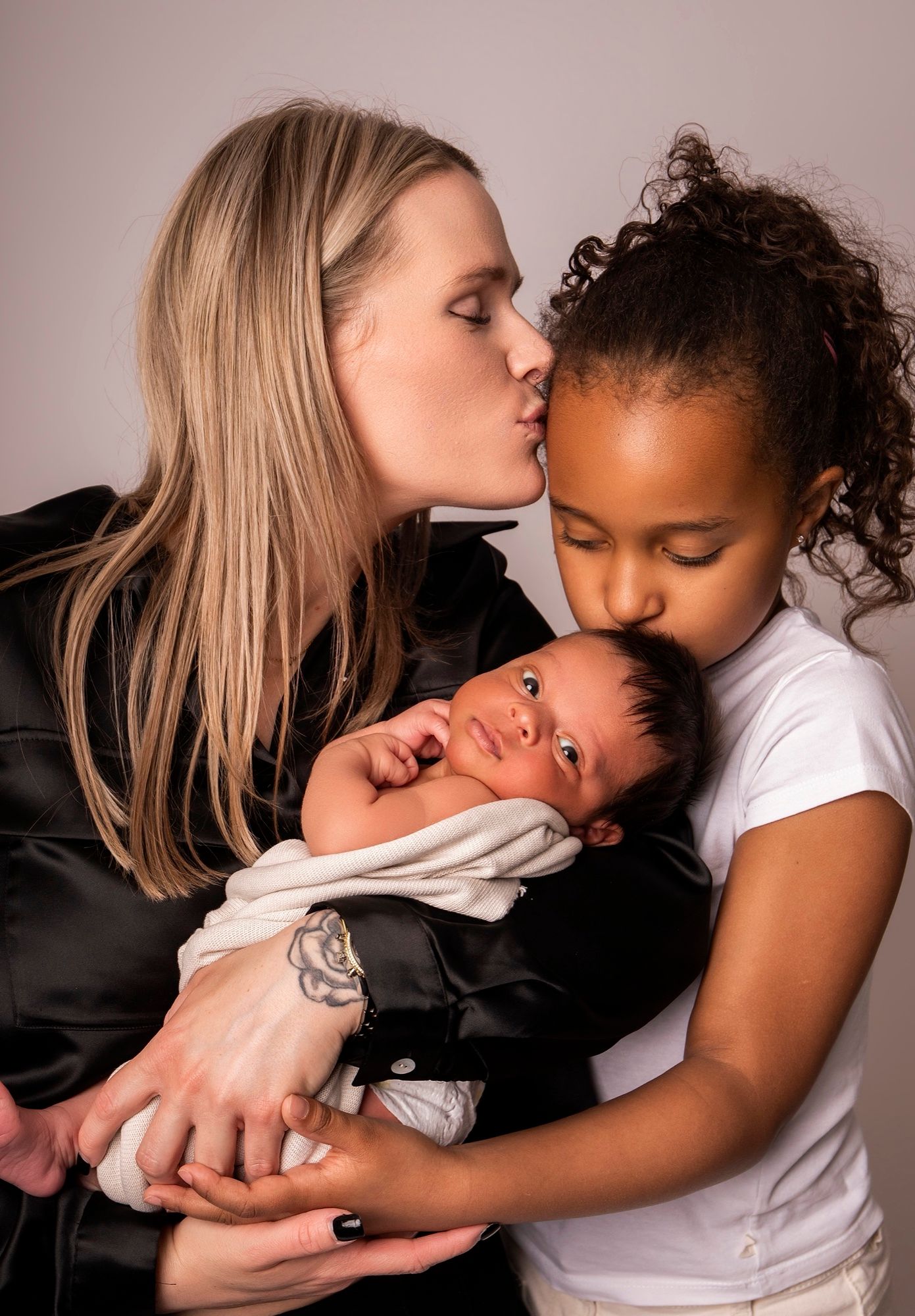 Mother kissing her older daughter, who in turn kisses the newborn in her arms, newborn photo session at the studio